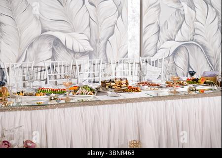 A beautifully set wedding reception table features an array of appetizers, cakes, and desserts. White chairs and a stylish backdrop enhance the elegan Stock Photo