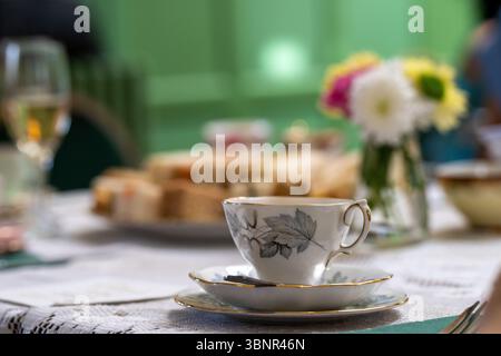 A small tea cup on a lace tablecloth with flowers and finger sandwiches in the background and a glass of champagne - high tea Stock Photo
