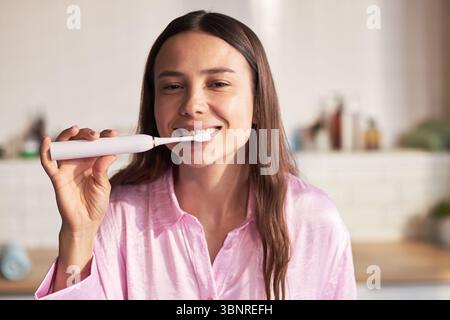 Portrait of young adult Caucasian woman smiling while brushing teeth with electric toothbrush in bathroom, demonstrating daily beauty and skincare routine for oral hygiene Stock Photo