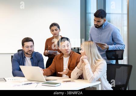 A diverse team collaborates around a table, discussing a laptop and sharing ideas. The focus is on teamwork and problem-solving. Stock Photo