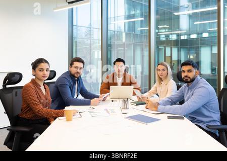 Diverse group of business professionals in a modern office setting, gathered around a table during a meeting, looking at the camera. Stock Photo