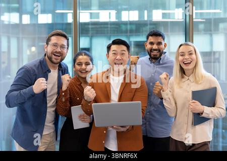 A diverse team celebrates a successful project with cheers and excitement, holding a laptop in a modern office environment. Stock Photo
