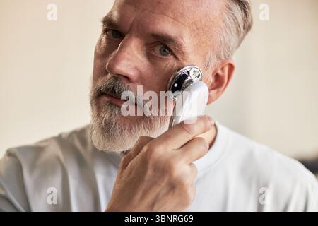 Portrait of middle aged Caucasian man using electric facial massager on cheek during skincare routine, maintaining eye contact with camera, demonstrating modern beauty technology Stock Photo