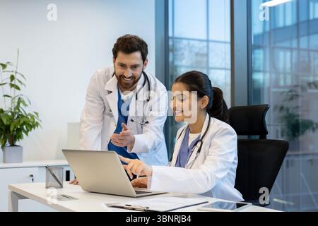 Two doctors, a man and a woman, collaborate at a desk in a modern office. They are looking at a laptop screen. Stock Photo