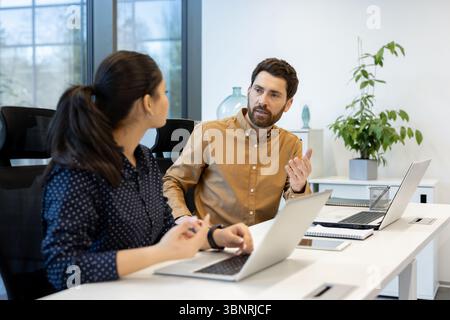 Two colleagues, a man and a woman, engage in a discussion in a modern office setting, working on laptops at a desk. Stock Photo