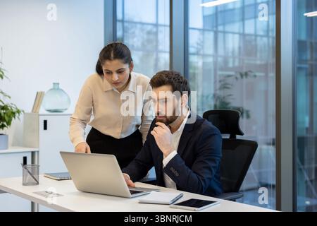 A diverse business team collaborates over a laptop in a modern office setting, discussing data and strategy. Stock Photo