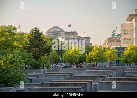 View of Berlin's Holocaust Memorial with Reichstag dome and Brandenburg Gate in background, during golden hour in summer. Stock Photo