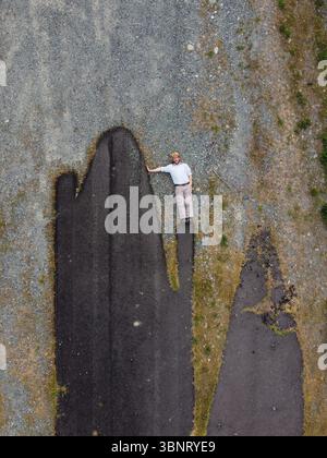Aerial view of a man standing on a dark, cracked asphalt surface amidst a sea of grey gravel and sparse green vegetation, Alessandria, Italy. Stock Photo