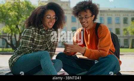 African American couple man guy male talking looking mobile phone social media showing smartphone discuss online game with smiling woman female girl Stock Photo