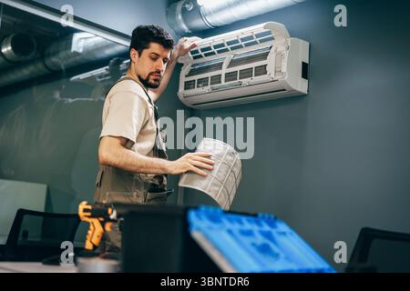 Technician performs air conditioner maintenance in modern office with blue walls and exposed ductwork Stock Photo