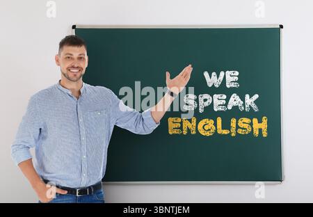 Teacher pointing at green chalkboard with text We Speak English. Language course Stock Photo