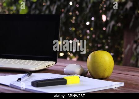 A laptop lying on a wooden surface outdoors. Next to the laptop is a yellow apple, as well as an open notebook with a pen and a yellow marker. Stock Photo