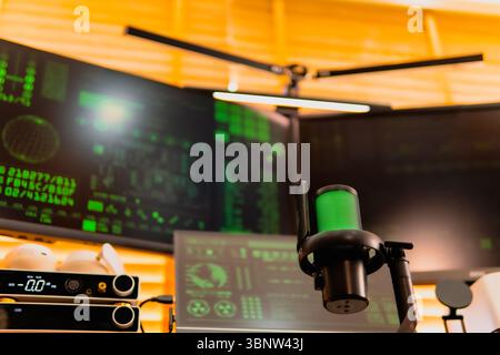 A close-up of a black microphone with green LED lighting, placed in front of multiple computer screens displaying hacker-style green codes and data. Stock Photo