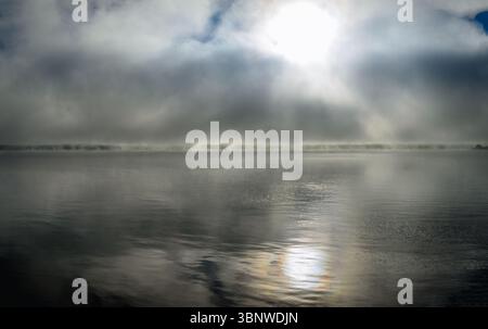 morning fog over the St. Lawrnence River showing the far coastline with light ripples in the water, and the sun reflecting on the near water. Stock Photo