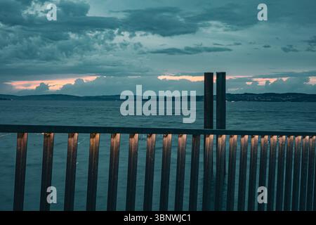 A calm evening view of Lake Constance with glowing lights shining through a metal fence. Mountains and clouds in the background create a moody atmosph Stock Photo