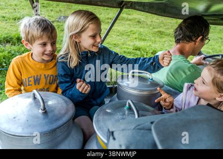Holiday experience in the Alps. Ruedi Zurfluh from Hobiel Alp takes guest children and freshly milked milk to the Alpine cheese dairy, Engelberg Valley at Surenen Pass. In Attinghausen, Switzerland, children and an adult are travelling in a vehicle transporting milk cans. They seem to be enjoying the journey together through the green countryside. Attinghausen, Uri, Switzerland Stock Photo