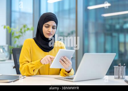 A professional Arab woman wearing a hijab is working at her desk in an office setting, using a tablet and laptop. Stock Photo