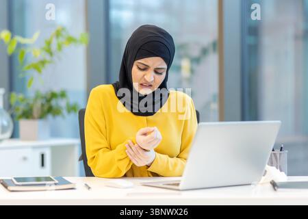 A woman wearing a hijab grimaces in pain, holding her wrist while working on her laptop in an office setting. Stock Photo