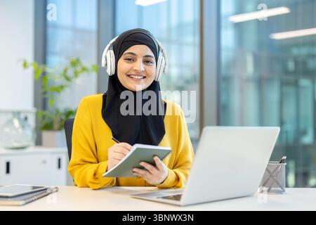 A smiling Muslim woman wearing a hijab and headphones, working on a laptop and taking notes at her desk in a modern office. Stock Photo