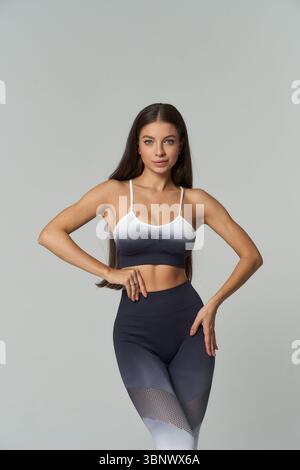 Young woman with long hair in sporty outfit poses confidently, wearing gradient top and leggings, hands on waist, displaying fit physique, neutral exp Stock Photo