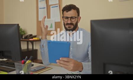 Hispanic man with beard in office looks puzzled holding blue tablet between dual monitors while wearing glasses and light blue shirt indoors. Stock Photo