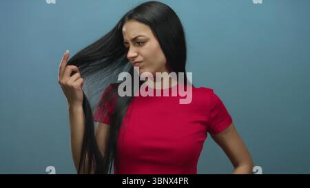 Woman with long hair in red shirt frowning and inspecting split ends against isolated blue background showing frustration. Stock Photo