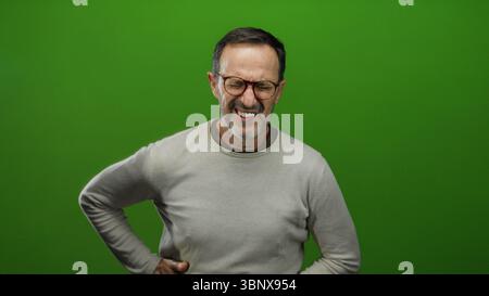 Man experiencing discomfort holding stomach over green background with glasses and gray sweater, suggesting pain or illness in middle-aged hispanic ma Stock Photo