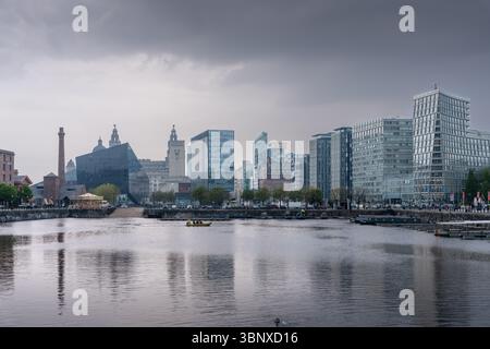 A panoramic scene of Liverpool’s Albert Dock at sunset under heavy clouds, showcasing a contrast between historic brick warehouses and modern glass bu Stock Photo