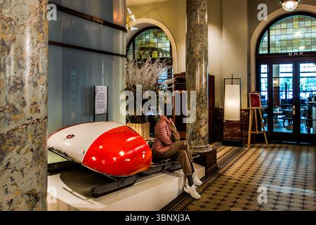 There is a historic bobsleigh in the entrance hall of the Kempinski Palace Engelberg. A mannequin sits in an elegant hotel lobby in Engelberg, Obwalden, Switzerland, next to a red bobsleigh bearing the Swiss cross and displayed on a pedestal. This scene at the Hotel Kempinskii emphasises the town's connection to winter sports Stock Photo