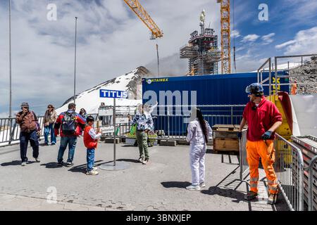 Indian tourists pose on the Titlis between construction cranes and construction workers. Tourists and a worker are seen on the summit of Mount Titlis in Engelberg, Obwalden, Switzerland. Visitors interact with a sign that reads 'I Love Switzerland' while construction work on a structure with cranes takes place in the background. The scene shows the mix of tourism and infrastructure development in this alpine region of Europe Stock Photo
