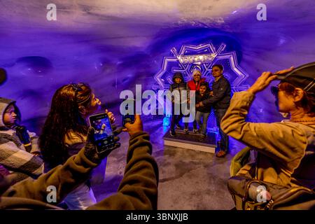 The Titlis Glacier Grotto is a tourist attraction at an altitude of over 3,000 metres, located directly at the Titlis cable car summit station. The grotto consists of an approximately 150-metre-long, artificially created passageway that leads around 20 metres below the surface of the glacier. The tunnel features photo spots such as a throne made of eternal ice. Visitors, including a family, pose and take photos of themselves in the impressive, blue-lit ice grotto on the Titlis in Engelberg, Obwalden, Switzerland. The 'TITLIS' ice sculpture serves as a striking backdrop for souvenir photos, whi Stock Photo