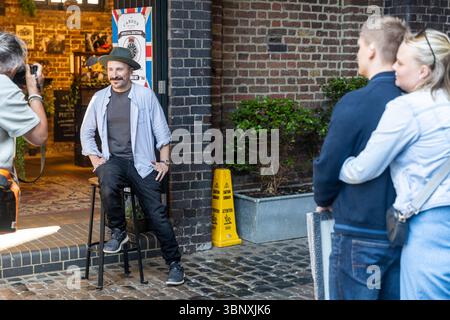London, UK, 11 May 2025,  A Photographer Captures a Portrait of a Man in a Hat Sitting on a Stool, with a Couple Nearby. Stock Photo