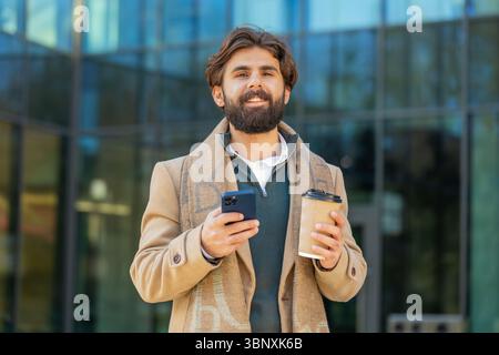 Happy Indian businessman enjoying morning coffee hot drink and using smartphone sending messages outdoors. Relaxing, taking a break. Man guy standing on urban city center street, drinking coffee to go Stock Photo