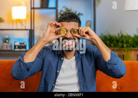 Portrait of smiling Indian freelancer man stock trader holding two golden bitcoins BTC. Happy Arabian male guy successful developer programmer sitting on sofa at home living room. Increasing wealth. Stock Photo