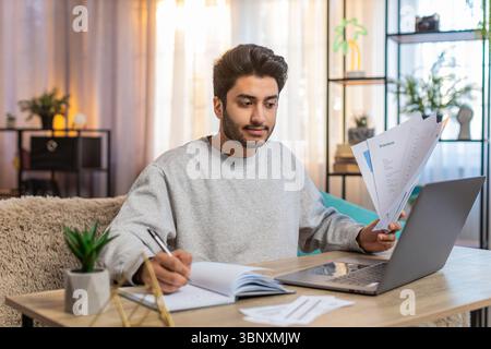Young Indian man freelancer sitting at table reviewing financial reports on laptop, holding documents and receipts, taking notes in notebook. Hispanic guy sits nearby at home, focused on his own task. Stock Photo