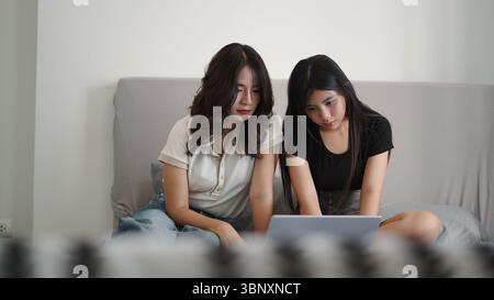 Two students are using a laptop, sitting on a bed, collaborating on a project Stock Photo