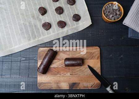 Flat lay of cookie dough bar on baking paper with knife cutting board and bowl of chocolate chips on black wooden background ready for baking Stock Photo