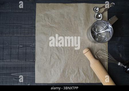 Top view of baking tools on dark wooden table with parchment paper stirring stick flour sieve and measuring spoons ready for baking Stock Photo