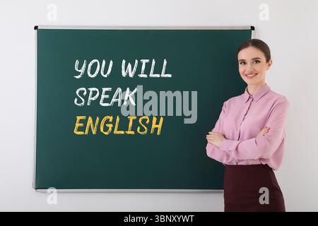 Teacher near green chalkboard with text You Will Speak English. Language course Stock Photo
