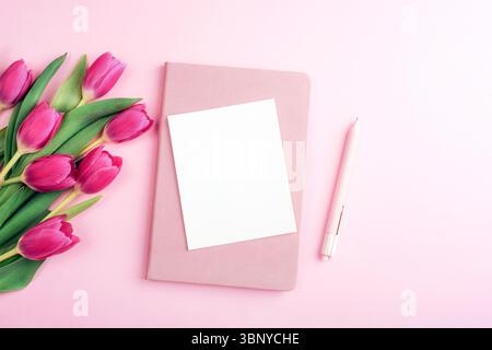 Blank white card with pink tulips, notebook and pen on pastel pink background. Top view, flat lay, mockup. Stock Photo
