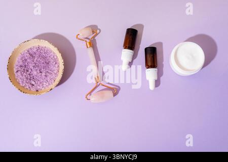 Spa cosmetic set with cream jar, essential oil bottles, rose quartz face roller and sea salt on lilac background with light and shadows. Top view, fla Stock Photo
