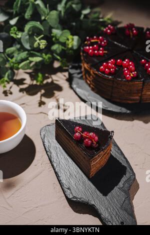 food still-life, A slice of rich chocolate cake garnished with fresh red berries rests on a dark slate platter. A cup of tea sits nearby, creating a w Stock Photo