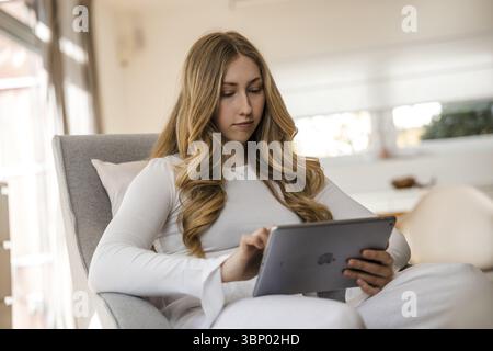 Young, blonde woman in a white top sits relaxed on an armchair with an iPad, concentrating Stock Photo