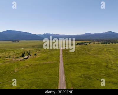High angle aerial view of an empty asphalt road stretching through vast green plains toward distant mountains near Crater Lake, Oregon. Stock Photo