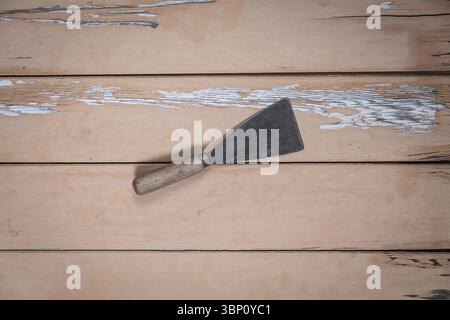 Old metal putty knife with wooden handle lying diagonally in the center of a worn beige wooden table, seen from a top-down perspective. Stock Photo