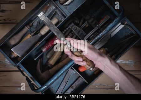 Hand holding an old chisel with wooden handle above an open blue metal toolbox filled with vintage tools on a worn wooden table. Stock Photo