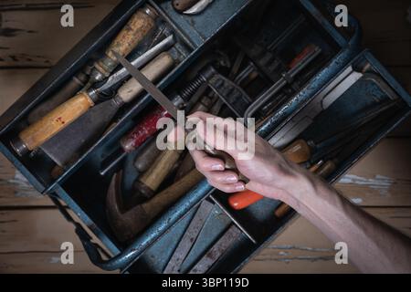Hand holding an old metal file above an open blue metal toolbox filled with vintage tools on a worn wooden table. Stock Photo