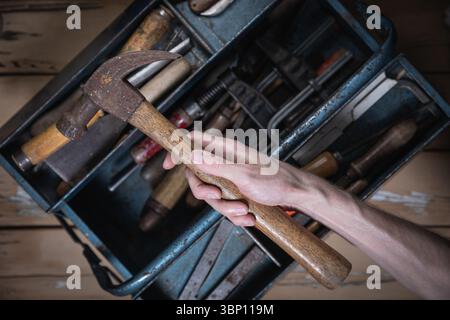Hand holding an old hammer with wooden handle above an open blue metal toolbox filled with vintage tools on a worn wooden table. Stock Photo