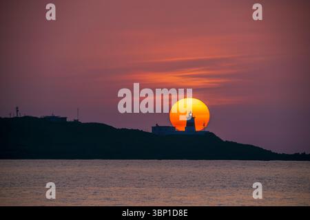 A breathtaking sunset view with the sun setting behind Fugui Cape Lighthouse, casting a warm glow over the sea in New Taipei City, Taiwan. Stock Photo