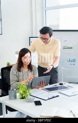 Two Asian business professionals review sustainability data. They discuss environmental social governance strategies. Office setting features renewabl Stock Photo